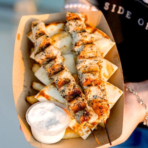 Two grilled chicken souvlaki skewers with pita bread and tzatziki sauce in a paper tray.